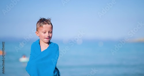Happy little boy wrapped in a blue towel, smiling joyfully on a sunny summer beach with the clear ocean in the background, enjoying a fun vacation by the water.