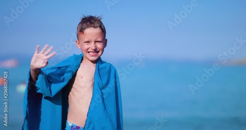 A joyful little boy on a bright summer day, wrapped in a blue towel, waves cheerfully at the camera with the beautiful blue sea behind him.
