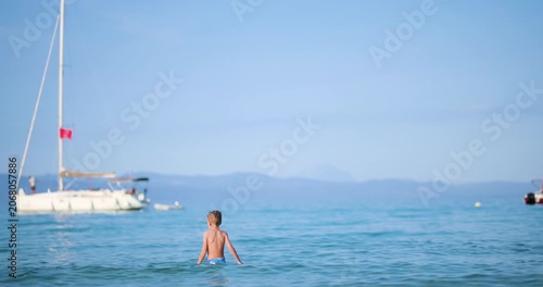 Adorable Young Boy Stands in Clear Blue Ocean Water, Gazing at a Distant Sailboat Under a Bright Summer Sky, Embracing the Freedom and Tranquility of Vacation.