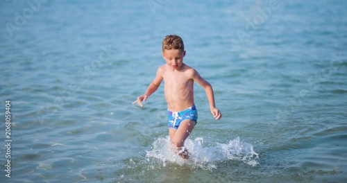 A happy little boy runs and splashes joyfully in the clear blue shallow water of the sea on a sunny summer day, enjoying his vacation at the beach.