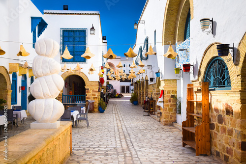 Принти на полотні Scenic narrow street with colorful flower pots and woven straw hats hanging above in the medina of Yasmine Hammamet, Tunisia