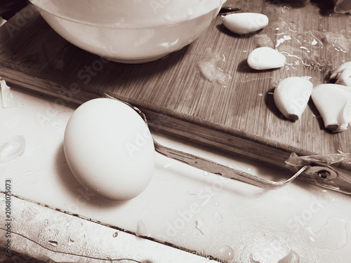 A sepia-toned close-up of a raw egg resting beside a cutting board with peeled garlic cloves during meal preparation. Water droplets, kitchen utensils, and a mixing bowl