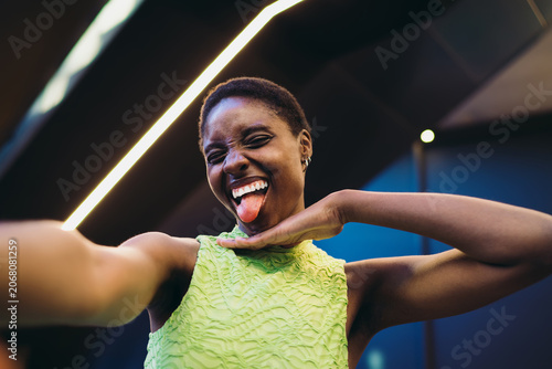 Obraz na plátne Excited black woman in neon dress showing tongue and capturing funny selfie on mobile phone