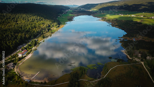 Aerial view of Lac des Rousses surrounded by lush green forests and rolling hills under a sky with scattered clouds in Les Rousses, Bourgogne-Franche-Comte, France.