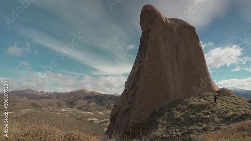 Hiker walking next to a gigantic vertical weathered stone monolith on a grassy hill, with a valley and hills under a blue sky with soft wispy clouds in La Buitrera, Bariloche, Argentina.
