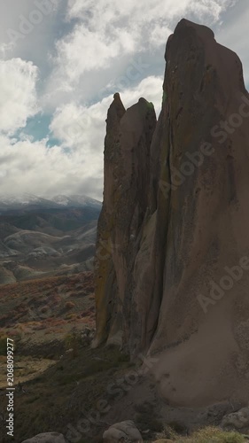 Giant vertical stone formations and rock spires rising over a valley with autumn vegetation and distant snow-capped mountains under a cloudy sky in La Buitrera, Bariloche, Argentina.