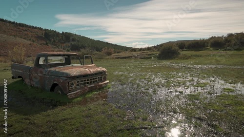 Wide shot of a vintage, rusted pickup truck abandoned in a swampy green field with puddles of water, surrounded by forested hills and bushes under a cloudy sky.