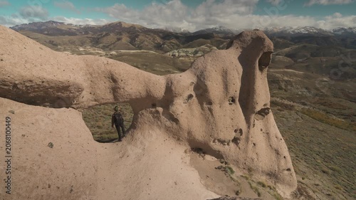 Wide shot from a high angle of a male hiker with a backpack standing inside a natural stone arch formation, looking out over a vast mountain valley under a cloudy sky in La Buitrera, Bariloche, Argent