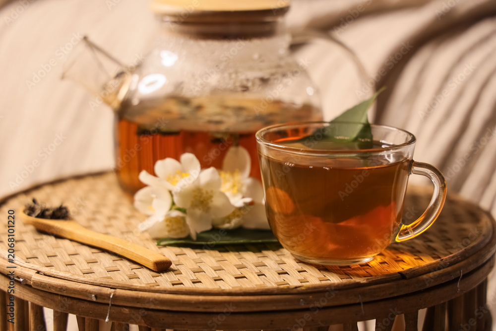 Naklejka premium Relaxing jasmine tea moment with glass teapot and cup on woven tray, cozy home comfort and wellness beverage, fresh jasmine flowers and a wooden spoon with tea leaves complete the tranquil composition