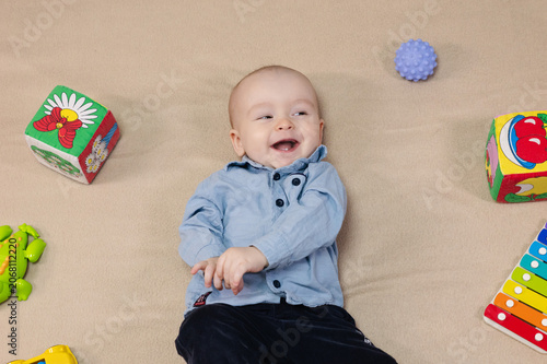 Cute 10 month baby boy lying among various playful colorful toys on beige blanket. Infant surrounded by vivid objects participating actively in sensory exploration and learning. 