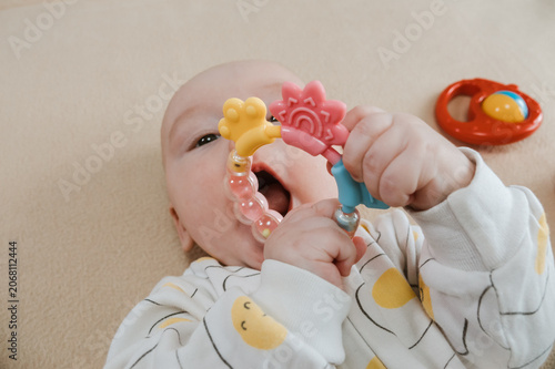 Baby lying on bed and gripping teething ring with both hands. Child chewing toy for teething relief. Exploring textures while soothing sore gums. Sensory discovery at home.