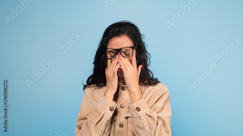 фотографія Worried Young Woman With Dark Hair, Rubbing Her Eyes With Hands, Displaying Signs Of Stress And Fatigue While Standing Against Solid Blue Background