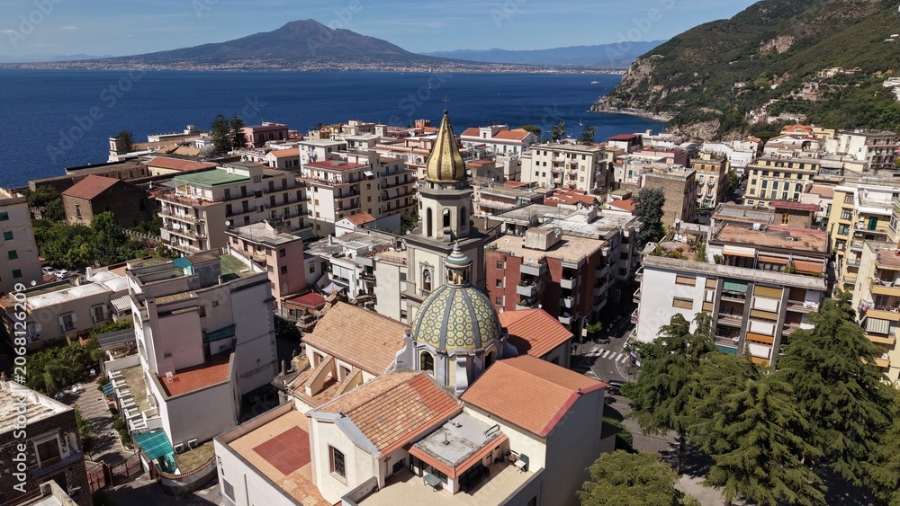 Fototapeta premium Aerial drone view of Vico Equense town with Chiesa dei Santi Ciro e Giovanni dome, terracotta rooftops and Mount Vesuvius across the gulf, Italy 