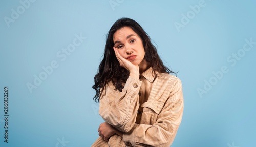 Foto Bored Hispanic Woman Resting Head On Hand, Looking Thoughtful While Standing With Arms Crossed In Studio Setting With Blue Background