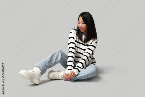 Beautiful young Asian woman in stylish jeans sitting on grey background