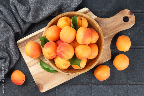 Ripe apricots with green leaves arranged in wooden bowl on rustic cutting board. Fresh seasonal harvest, summer fruit on black tile background healthy eating, food ingredient for cooking and desserts