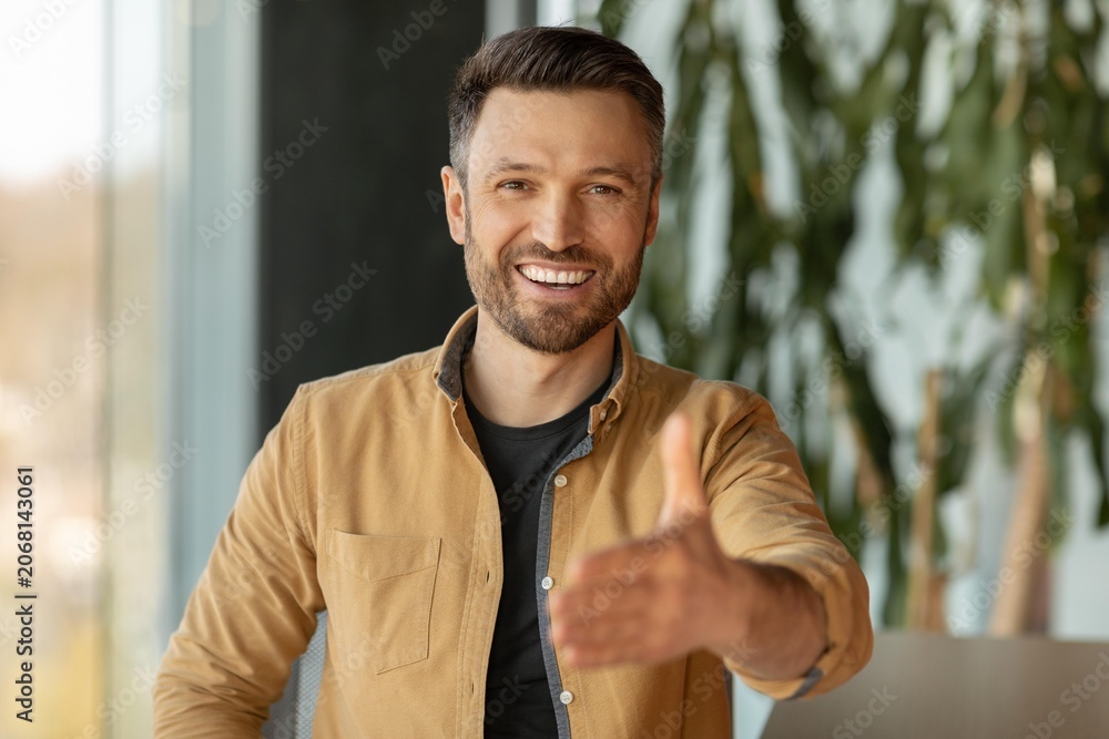 Obraz premium Smiling Caucasian Man In His 30s Giving Handshake While Sitting At Office, Looking Confident And Engaged, Representing Business Interaction And Agreement, Professional Setting.