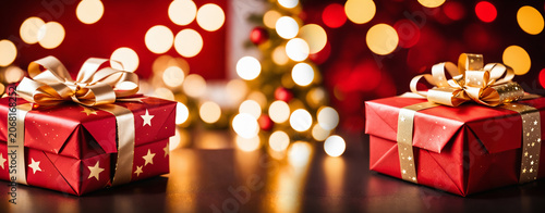 two red gift boxes decorated with golden stars and ribbons resting on a dark surface against a blurred christmas tree with warm glowing bokeh lights.