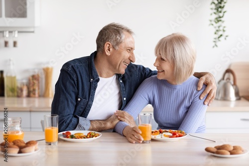 Smiling Elderly Caucasian Man And Woman Having Breakfast Together In Kitchen  Enjoying Healthy Meal Of Fruits And Vegetables  Sitting At Table With Drinks And Laughing. Happy Moment.