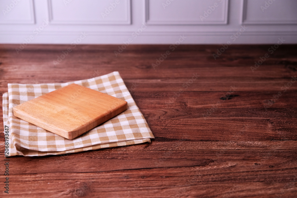 Fototapeta premium Square wooden board rests on checkered cloth. Wood grain shows warm, natural texture. Dark wood table provides rustic backdrop. Simple, clean composition invites creativity
