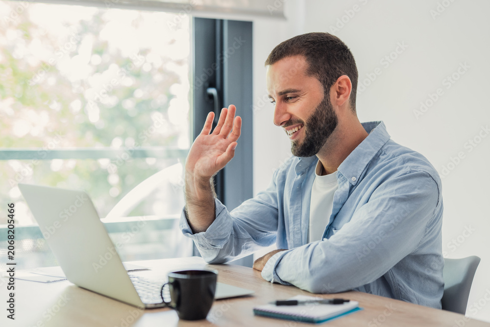 Obraz premium Man smiling and waving to a laptop screen while participating in an online video call from his bright home office, connecting remotely