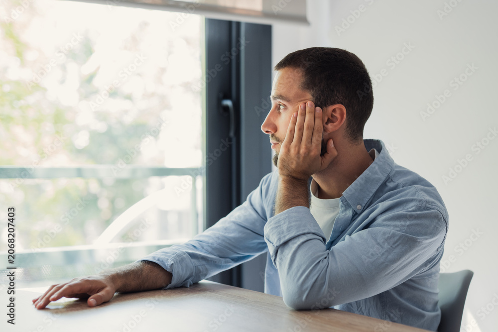 Obraz premium Young man sitting at a table at home. Thoughtfully looking out the window. Embracing introspection as he considers his future plans or past events in a moment of quiet reflection and mental processing