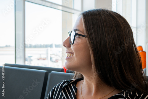 фотографія Smiling woman wearing glasses sitting in an airport terminal, looking out of the