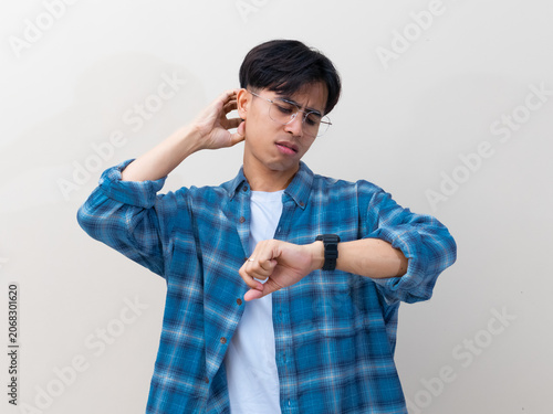 Fotografie Confused young Asian man with glasses looking at his wristwatch while scratching