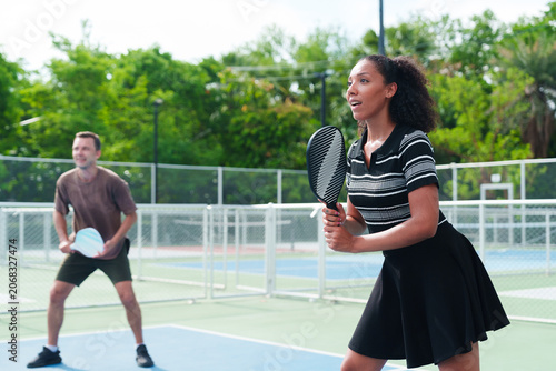 Diverse team of black woman and white man playing pickleball, looking at the opponent on modern outdoor court on sunny day. Sports, active and healthy lifestyle concept
