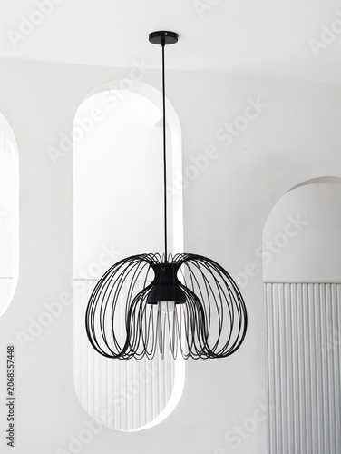 Fotografi A minimalist modern pendant light fixture with a black wire-frame design, suspended in a bright interior room featuring white arched architectural elements