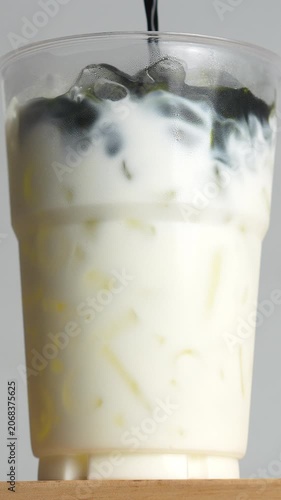 4K Vertical shot of iced matcha green tea latte in clear plastic glass on wooden table, Delicious layered beverage with rich green tea pouring into fresh white milk.