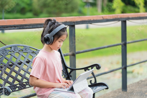 Young girl sitting on park bench wearing headphones and using tablet device outdoors with greenery background and metal fence for relaxing and entertainment