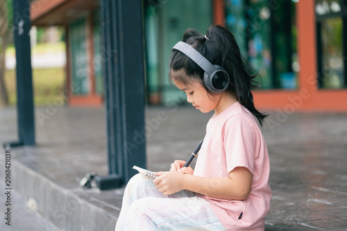 Young girl sitting outdoors on a bench, wearing headphones and writing in a notebook, enjoying music and exploring creativity in a vibrant urban park setting
