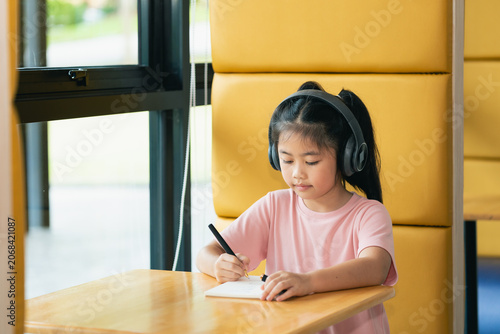 Young girl sitting at wooden table wearing large black headphones, writing in notebook, in bright modern space with large window, yellow wall, and natural light