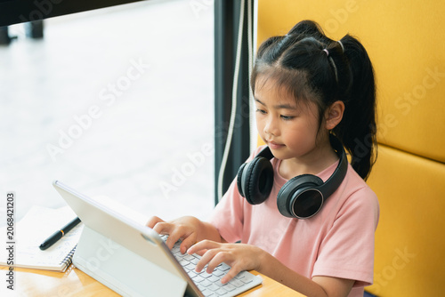 Young girl with dark hair in ponytails using a tablet device while sitting in a modern cafe with yellow seating, wearing a pink shirt and headphones, in a bright and casual environment
