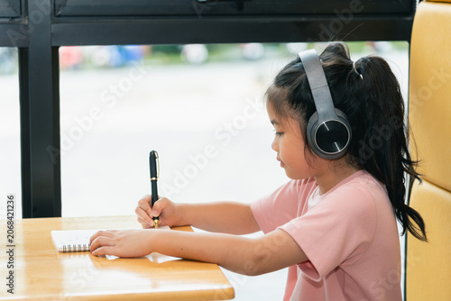 Young girl wearing large headphones sitting at wooden table writing in notebook with pen, indoor setting, focused expression, natural light, casual clothing, education concept