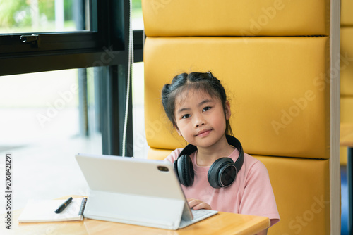 Young Girl Sitting at Desk Using Tablet with Headphones on in Modern Classroom Setting Near Window and Yellow Antique Wall Panel