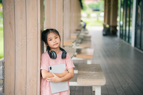 Young Asian Girl Holding Book and Notebook in Modern Indoor Space with Wooden Walls and Benches, Casual Clothing, and Headphones, Expressing Thoughtfulness and Curiosity