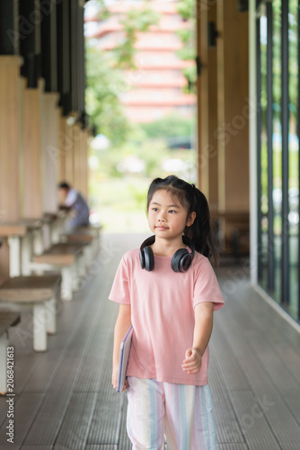 Young Asian girl with long dark hair, wearing pink t-shirt and colorful striped pants, standing on modern outdoor corridor with headphones around her neck, holding a notebook