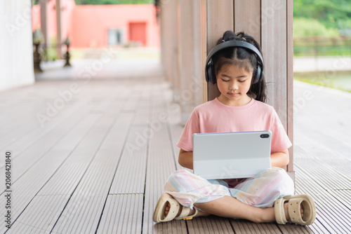 Young girl sitting cross-legged on wooden deck outdoors, wearing headphones and holding tablet device, enjoying digital entertainment in natural setting