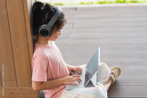 Young girl relaxing outdoors sitting against wooden wall, using laptop, wearing large headphones, casual clothing, enjoying digital entertainment on patio