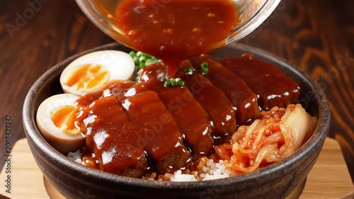 Close-up of thick dark red sauce being poured over sliced braised pork belly served with soft boiled eggs, kimchi and green onions on white rice in a dark ceramic bowl on wood.