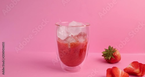 Dropping cold ice cubes into fresh strawberry smoothie glass on pink background. Refreshing summer beverage concept showcases dynamic motion blur with organic red berries on colorful surface