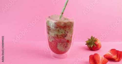 Stirring layered strawberry matcha latte on pink background. Female hand mixes refreshing iced green tea cocktail with organic red berry puree inside double walled glass. Detailed lifestyle close-up