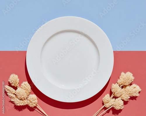 Round white ceramic plate mockup on blue and coral background with dried wheat stalks for seasonal menu design, autumn holiday dinner table setting, and food branding