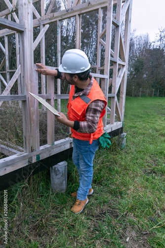 Construction worker inspecting a wooden house frame using a digital tablet on-site.