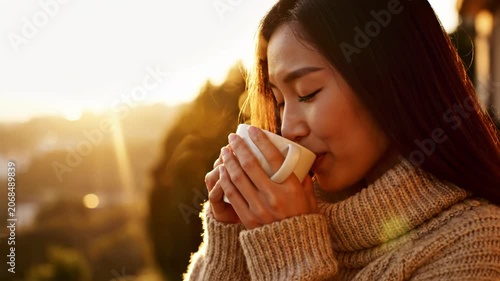 Young woman with long dark hair wearing a beige knit turtleneck sweater, smiling and holding a steaming white ceramic mug during a golden hour sunset with soft background light.