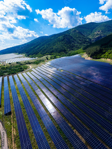 Solar panels cover fields in a mountainous area during a bright day with few clouds and sunlight shining on the solar arrays