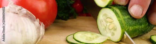 Preparing a fresh salad a chef is slicing a cucumber with a knife on a wooden board next to other vegetables.