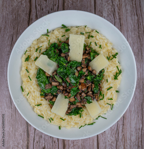 A bowl contains a serving of mushroom and spinach risotto. On top are pieces of Morteau sausage and parmesan cheese. The dish is placed on a wooden surface.